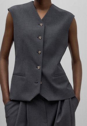 Ärmellose maßgeschneiderte dunkelgraue Weste mit Knöpfen vorne und Händen in den Taschen passender plissierter Hosen vor neutralem Hintergrund.