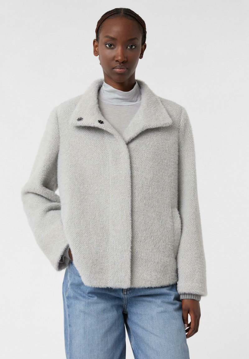 Giacca grigia chiara con texture, colletto, due bottoni sul collo e tasche laterali, abbinata a una maglia grigia e jeans blu.