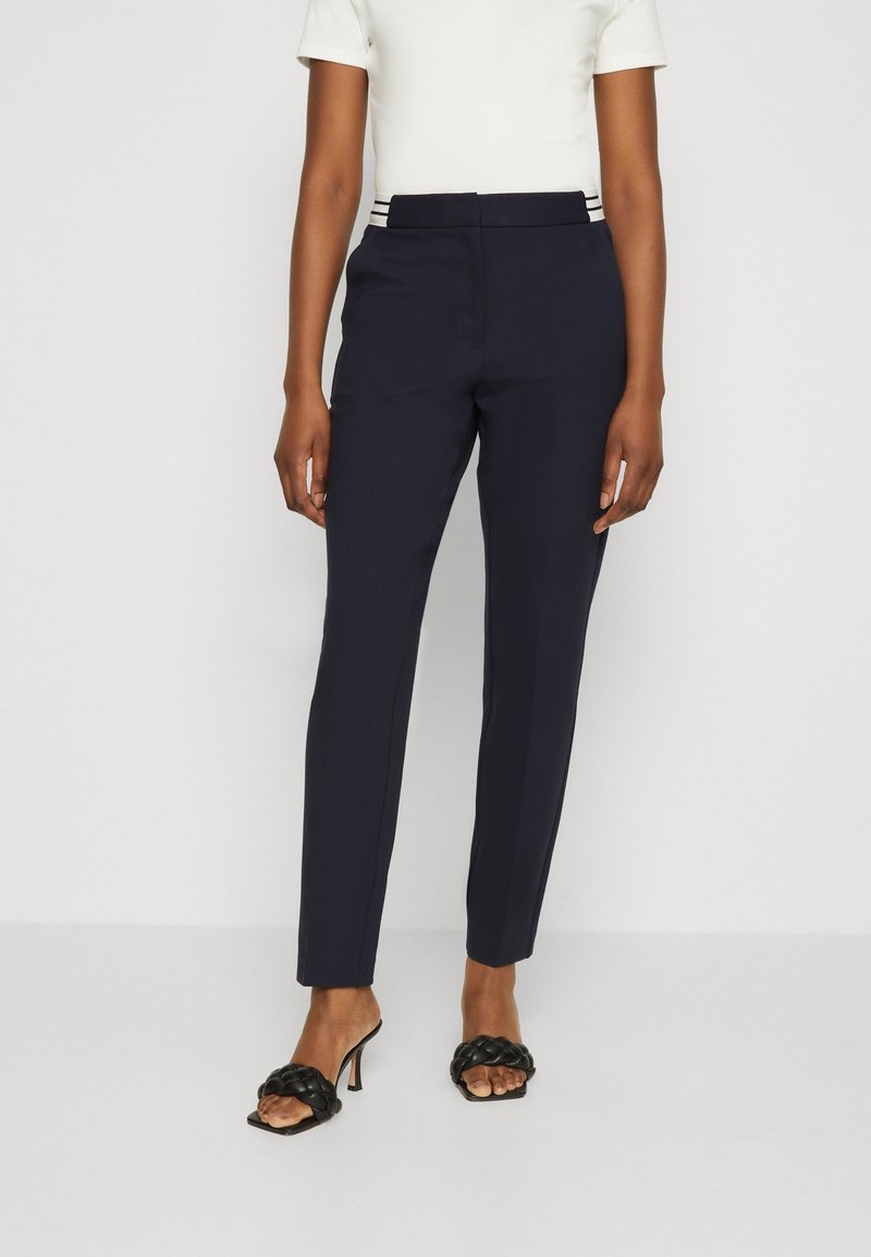 Personne portant un pantalon fuselé bleu marine taille haute, un haut blanc à manches courtes et des sandales à talons ouvertes noires tressées, debout contre un mur blanc.