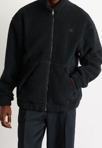 Chaqueta de forro polar negra con cuello alto, cierre frontal y dos bolsillos laterales. Presenta un tejido texturizado y un sutil logo de la marca en el pecho.