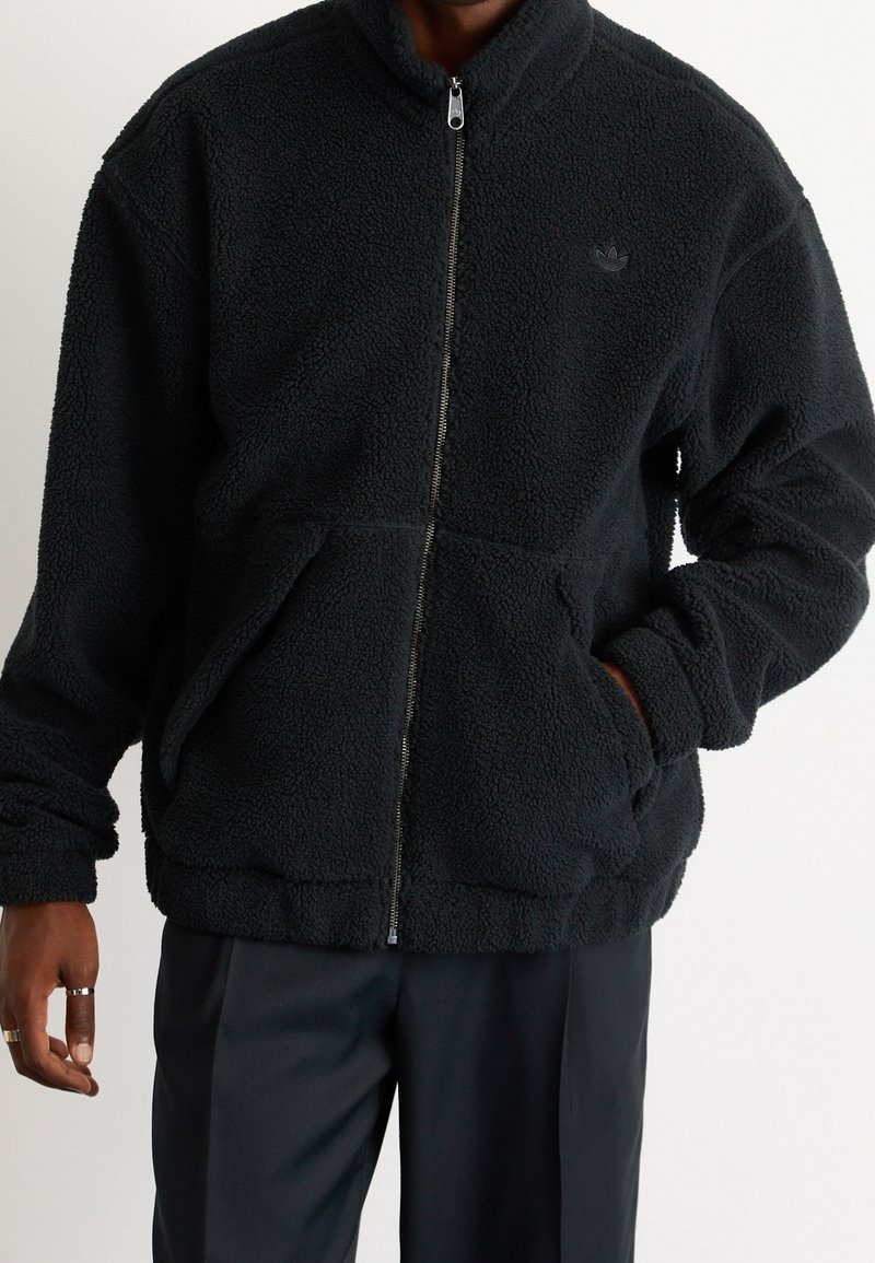Chaqueta de forro polar negra con cuello alto, cierre frontal y dos bolsillos laterales. Presenta un tejido texturizado y un sutil logo de la marca en el pecho.