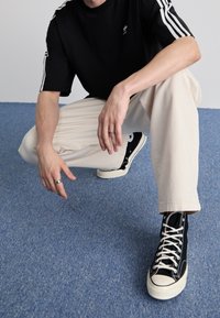 Svart atletisk tröja med vita ränder på ärmarna, beige byxor och svarta högklackade sneakers med vit sula och snören, sittande på knä på en blå matta.