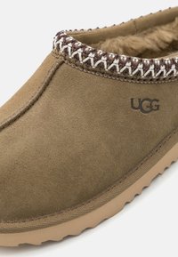 Brun mocka slip-on-sko med vit och brun dekorativ sömnad vid kragen, med en mjuk inredning och en strukturerad gummisula.