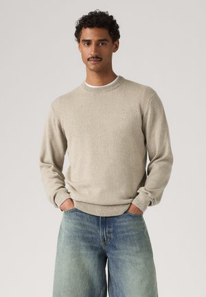 Uomo con capelli ricci e baffi, che indossa un maglione beige e jeans blu ampi, in piedi con le mani nelle tasche davanti a uno sfondo semplice.