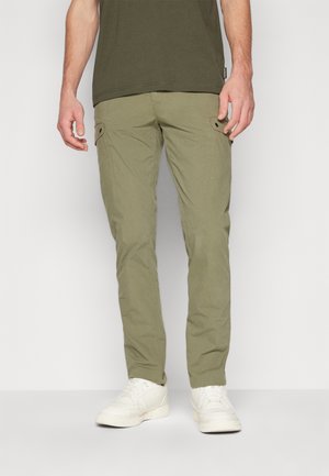 Mann trägt olivgrüne Cargo-Hose und weiße Sneakers, steht vor einem schlichten hellen Hintergrund.