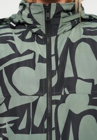 Πράσινο και μαύρο μπουφάν με μοτίβο, ψηλό γιακά και κλείσιμο με φερμουάρ. Το υλικό φαίνεται λείο με αφηρημένα μαύρα σχέδια σε αμυδρό πράσινο φόντο.