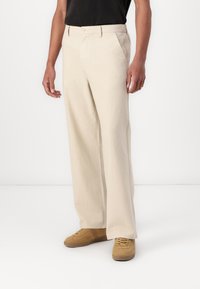 Bredde bukser i beige laget av et strukturert stoff, med knappeåpning, sidelommer og en skreddersydd passform. Brukt med brune sneakers.