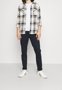 Camicia flanella a quadri in crema, blu e arancione, con bottoni e tasche, abbinata a pantaloni blu e sneakers bianche con dettagli grigi.