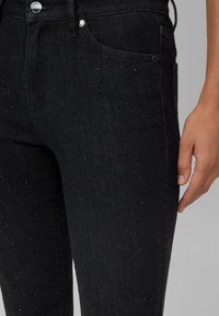Jean en denim noir avec une coupe ajustée, des détails légèrement usés subtils et une fermeture à boutons brillante. Texture lisse avec un léger éclat.