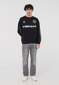 Felpa nera con la scritta "VIBRANT" in lettere bianche, dotata di una toppa rotonda e strisce bianche. Abbinata a jeans grigi chiari e sneakers bianche.