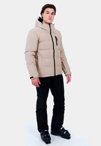 Chaqueta acolchada beige con capucha, cierre frontal y detalles en negro. El modelo lleva pantalones negros y botas de esquí, de pie frente a un fondo blanco.