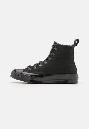 Musta korkeaankapenevat tennarit, jotka on valmistettu mokasta ja kiiltävästä lakatusta nahasta, joissa on teksturoitu viimeistely ja metalliset nepparit nauhoille.