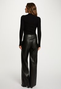 Donna in piedi di spalle, che indossa un top lungo a maniche nere a coste e pantaloni larghi in pelle nera a vita alta, su uno sfondo neutro.
