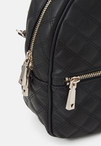 Sac à dos en cuir noir avec un motif matelassé, fermeture éclair dorée et détails de tirette géométriques. Design compact et arrondi avec deux compartiments zippés.