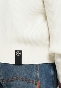 Deteljni prikaz donjeg ruba bijelog žbukanog džempera s crnom oznakom AX iznad džepa plavih traperica s narančastim šavom.