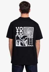 Man som bär en svart t-shirt med en vit grafisk design som har ordet "VOID", ett spiralformat mönster, en stege och abstrakta former.