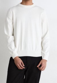 Weißer Strickpullover mit Rundhalsausschnitt, gerippten Bündchen und Saum sowie besticktem Logo auf der Brust. Getragen mit schwarzen Hosen.