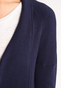 Tmavě modrý svetr se vzorovaným úpletem, s velk ým klopovým límcem a volným střihem, nošený přes světle šedé tričko.