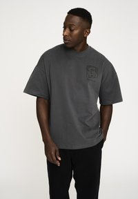 Grå kortærmet t-shirt lavet af bomuld. Har et frontlommeprint med et abstrakt ansigtsdesign og ordet "Resterind."