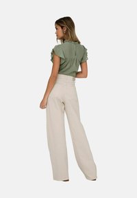 Blouse verte à manches froncées, dos en clé, associée à un pantalon large beige taille haute, affichant une texture lisse et douce.