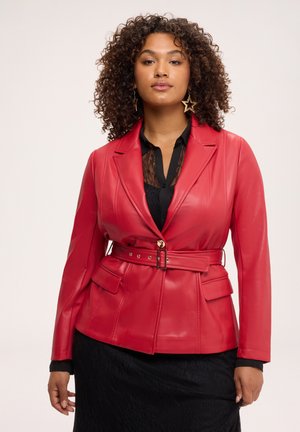 Donna con capelli ricci che indossa una giacca di pelle rossa con cintura sopra un top in pizzo nero e gonna nera, con orecchini a forma di stella.
