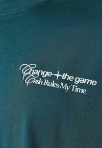 Tissu vert foncé avec texte stylisé blanc indiquant "Change the game", surmonté par le cursif "Cash" en haut et "Rules My Time" en bas.