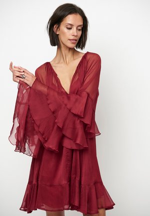 Vrouw draagt een losse, dieprode jurk met doorschijnende, gelaagde klokmouwen en een gerimpelde zoom, poseert met handen samengevouwen vlak bij de borst.