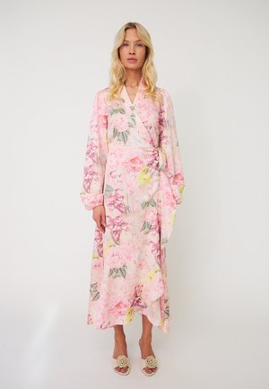 Femme blonde portant une longue robe portefeuille fleurie rose à manches longues et des talons blancs ouverts avec des détails floraux, debout devant un fond uni.