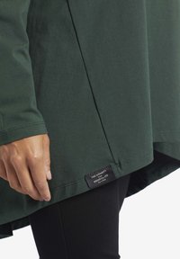 Tunic verde scuro con una texture liscia, orlo con spacco laterale e etichetta del marchio visibile. Caratterizzato da maniche lunghe e una vestibilità ampia per il massimo comfort.