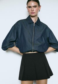 Denim-Cropped-Jacke mit Reißverschluss vorne, weiten Ärmeln und in Dunkelblau, kombiniert mit einem schwarzen, plissierten Minirock.