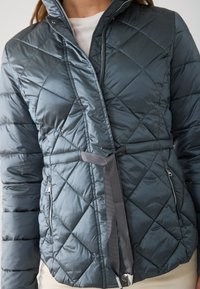 Tamsiai mėlynas, dvigubas pūkų kailis, turintis užtrauktuką, diržą juosmeniui ir užtraukiamas šonines kišenes papildomam funkcionalumui.