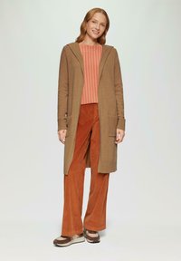 Brauner gestrickter Cardigan mit Kapuze, gestreiftes oranges Oberteil und rostfarbene Cordhosen. Die Schuhe sind beige mit braunen Akzenten und einer dicken Sohle.