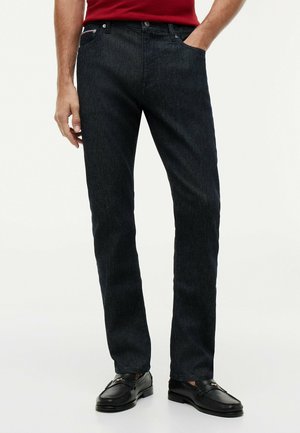 Mann trägt dunkelblaue Jeans mit geradem Bein, rotes Hemd und schwarze Leder-Loafers, steht mit einer Hand in der Tasche.