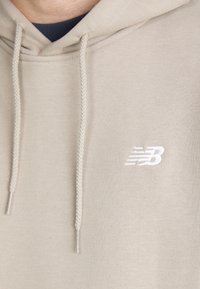 Felpa beige chiaro con cappuccio e cordoncino, realizzata in un tessuto morbido. Presenta un logo ricamato bianco sul petto.