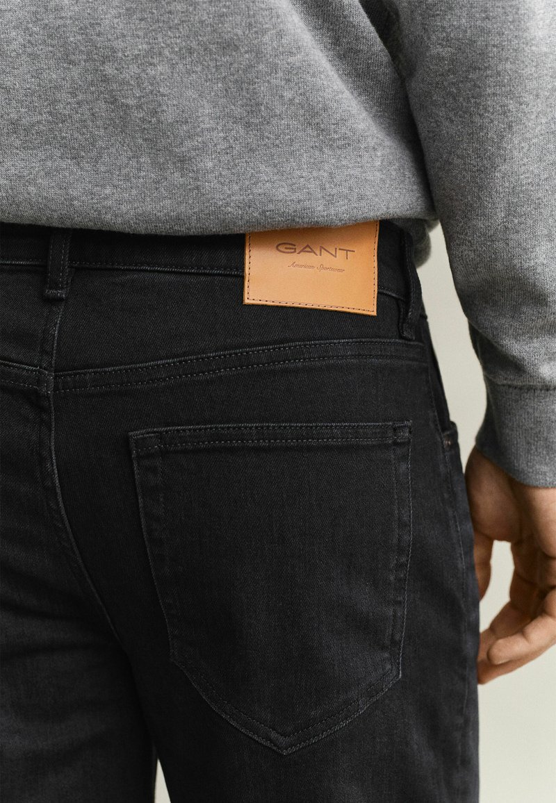 Vue de dos d'une personne portant un jean noir avec un patch en cuir marron de la marque GANT et un sweatshirt gris.
