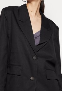 Sort blazer lavet af tekstureret stof. Har en spids krave, to forreste lommer og to sorte knapper til lukning. Laget over en lilla top.