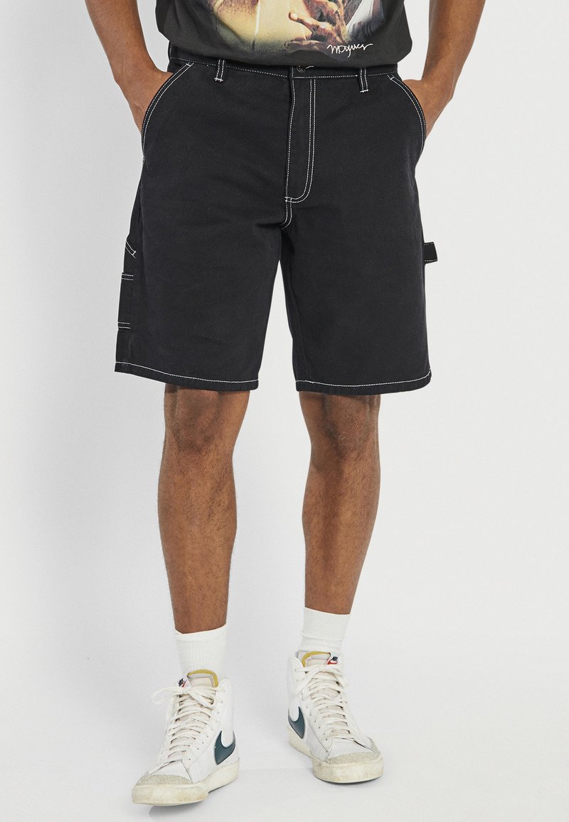 Black shorts made of cotton, featuring contrast white stitching, pockets on the sides, and a straight cut. Styled with white sneakers.