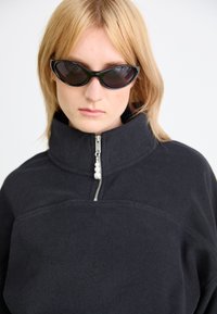 Schwarze Fleece-Zipperjacke mit hohem Kragen und perlenförmigem Reißverschluss. Das Model trägt schwarze, ovale Sonnenbrillen und hat glattes, blondes Haar, das das Gesicht umrahmt.