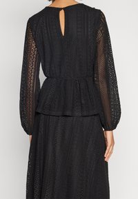 Abito nero con un design in pizzo testurizzato, maniche lunghe e vita a peplo. La parte posteriore presenta un'apertura a chiave e una chiusura con bottone.