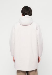 Manteau long, rose clair avec une capuche, présentant un design simple et une texture lisse. L'arrière est dépouillé, sans quincaillerie visible.