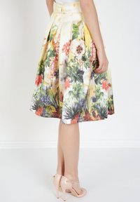 Falda con estampado floral que presenta un diseño plisado, predominantemente amarilla con flores verdes, rojas y moradas, combinada con tacones nude.