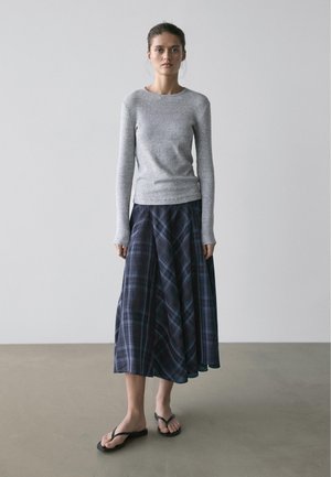 Frau steht barfuß auf grauem Boden, trägt ein graues Langarmshirt und einen blauen karierten Midirock vor einer hellgrauen Wand.