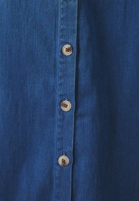 Denim srajca v temno modri barvi, z gumbom spredaj, ki vključuje šest svetlo bež gumbov, ter teksturirano tkanino in ravno krojenje.