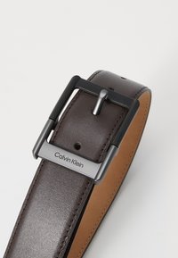 Barna bőr öv textúrált fekete fémből készült csattal. Ezüst színű, gravírozott "Calvin Klein" logóval és öt lyukkal a méretre állításhoz.