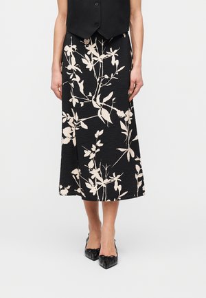 Kobieta ubrana w czarną spódnicę midi w beżowy kwiatowy wzór, czarny bezrękawnik oraz czarne buty na obcasie z ostrym noskiem, stojąca na białym tle.