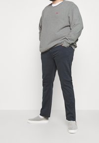 Homme portant un sweat-shirt gris, un sous-vêtement blanc, un pantalon bleu foncé et des chaussures grises sans lacets, debout les mains dans les poches contre un fond blanc.