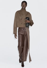 Brown double-breasted cropped wool coat paired with brown leather wide-leg pants and black textured clutch. Long sleeves and sleek, smooth texture.