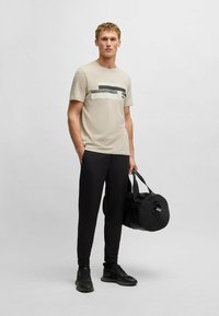 Camiseta beige con rayas horizontales en blanco y negro, combinada con pantalones negros y zapatillas negras, sosteniendo una bolsa de deporte negra.