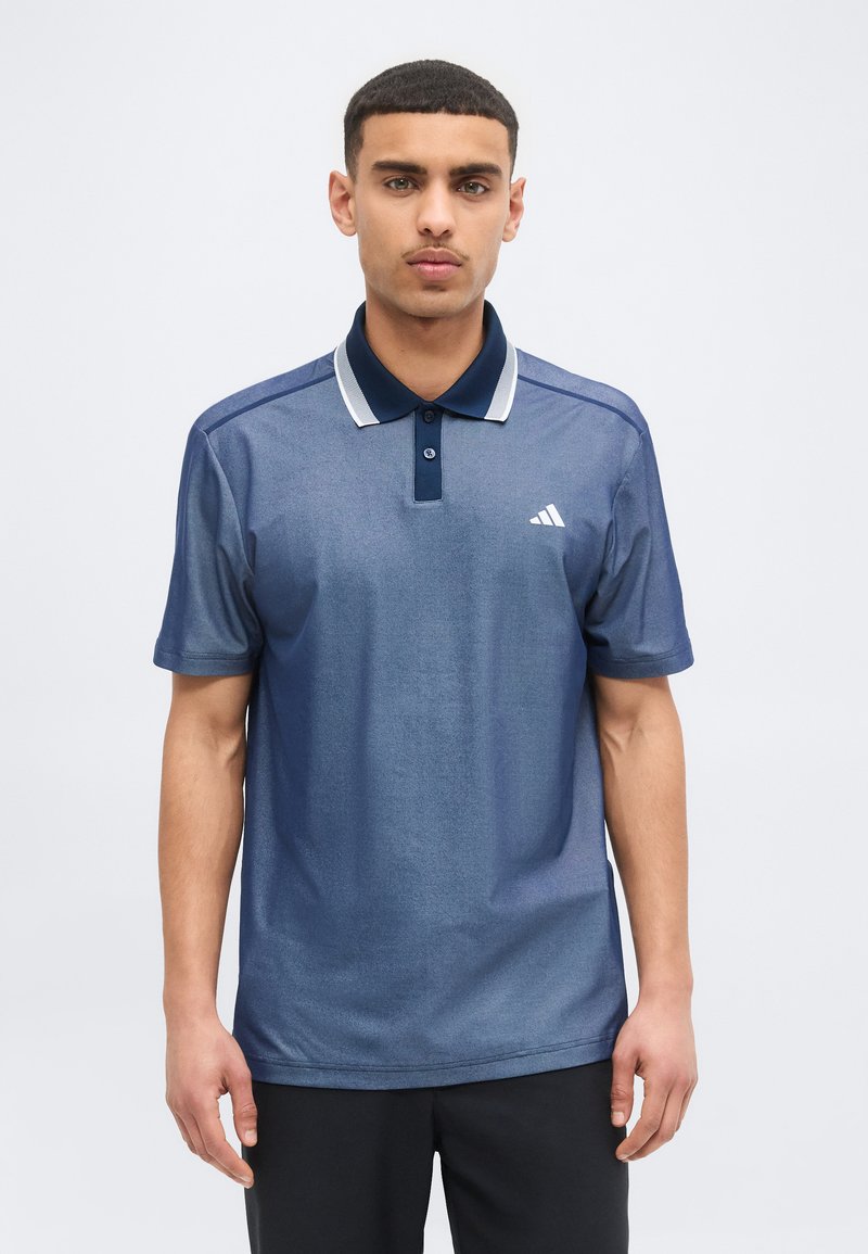 Νεαρός άνδρας με κοντά σκούρα μαλλιά, φορώντας μπλε πόλο μπλούζα Adidas και μαύρο παντελόνι, στέκεται μπροστά σε απλό ανοιχτόχρωμο φόντο.