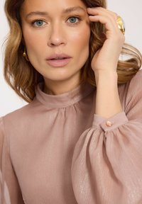 Kobieta z falującymi, jasnobrązowymi włosami, ubrana w błyszczącą różową bluzkę z bufkami, dotykająca twarzy, nosząca złote kolczyki i pierścionek.